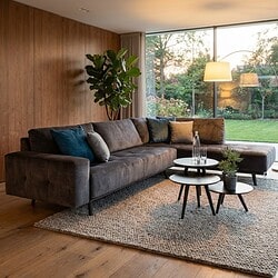 Elegante stoffen hoekbank in een moderne woonkamer met groot raam, natuurlijke lichtinval, houten wand en trendy bijzettafels. Perfect voor comfortabel en stijlvol wonen.