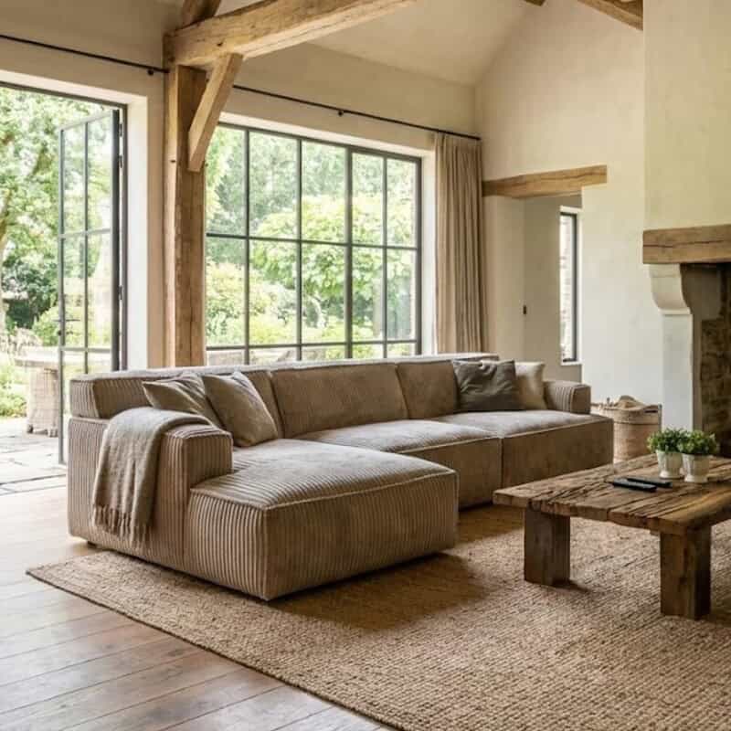 Luxe landelijke woonkamer met grote raampartijen, comfortabele beige stoffen hoekbank, rustieke houten salontafel en natuurlijke decoratie, perfect voor een gezellige en stijlvolle sfeer.