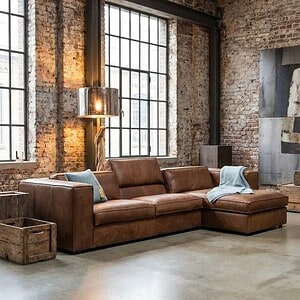 Elegante leren hoekbank in industrieel interieur met grote ramen en exposed bakstenen muren, perfect voor moderne woonkamers en lofts.