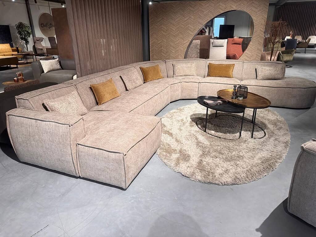 Luxe ronde modular bank in zachte beige stof met kussens, gecombineerd met een zwart metalen salontafel en een ronde crème vloerkleed in een modern interieur.