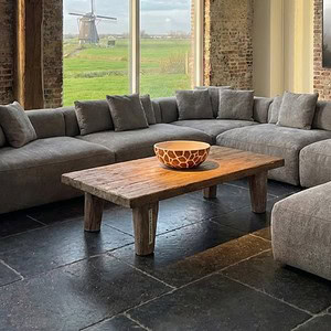 Houten salontafel in industriële woonruimte met grote grijze bank en uitzicht op typisch Nederlandse landschap met windmolen.