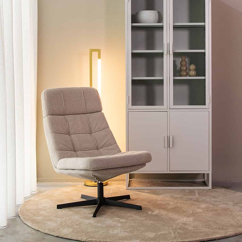 Minimalistisch design lounge fauteuil in zachte beige stof met zwart draaipoot, naast witte houten vitrinekast met glazen deuren, sfeervol interieur met warme verlichting en beige vloerkleed, perfect voor woonkamer.