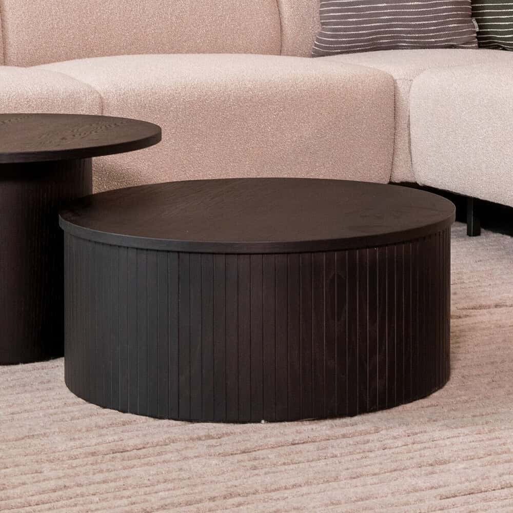 Grote ronde zwarte salontafel van Mokana Meubelen, trendy en functioneel voor moderne woonkamers, gecombineerd met een lichte bank en decoratieve kussens.