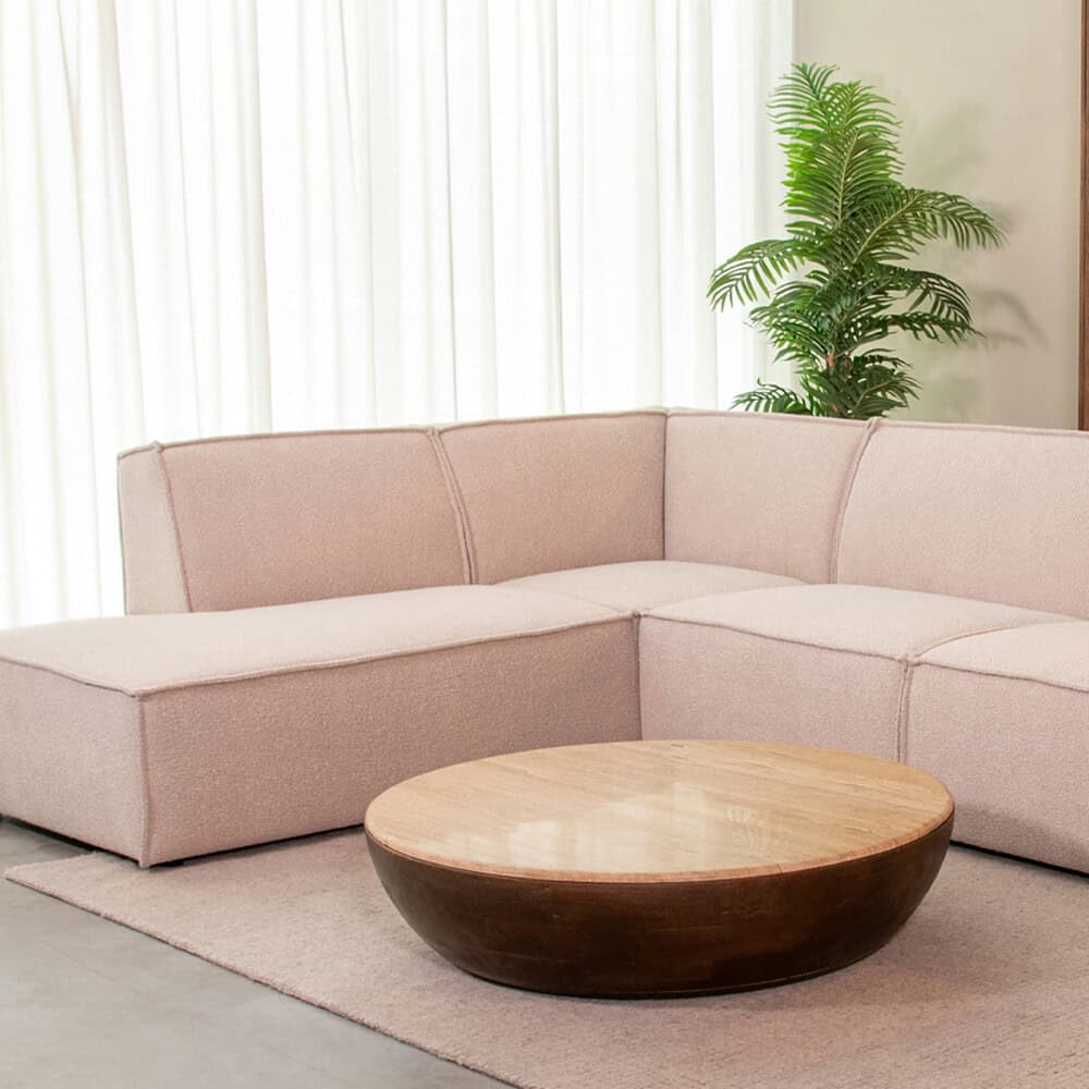 Elegante blushroze stoffen hoekbank met strakke lijnen, perfect voor modern interieur, gecombineerd met een ronde houten salontafel en groene planten voor een gezellige sfeer.