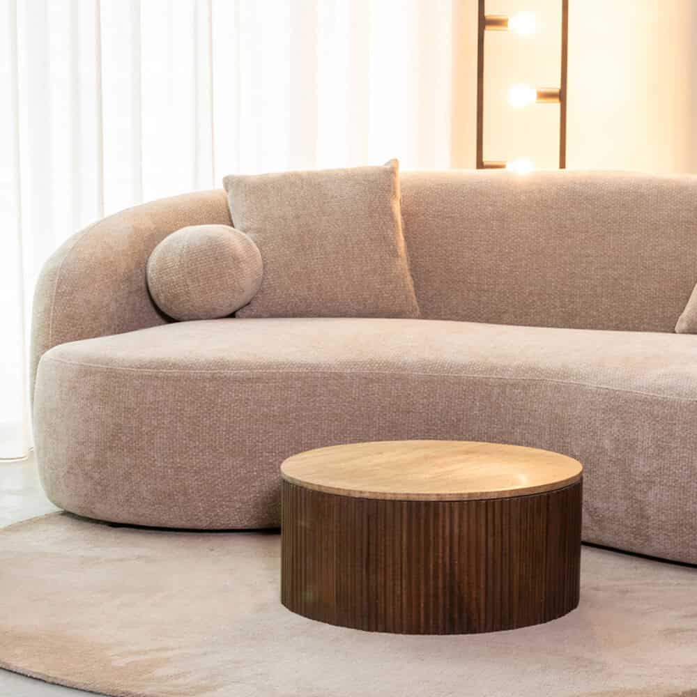 Sfeervol beige stoffen bank met ronde armleuningen en sierkussens, gecombineerd met een ronde houten salontafel in een modern interieur. Perfect voor een stijlvolle en gezellige woonkamer.