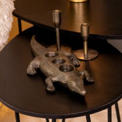 Chuck the high-res product photo showing a black circular table with a unique crocodile-shaped candelabra and two cylinder candles. The scene highlights modern, stylish home decor and furniture pieces from Mokana Meubelen.