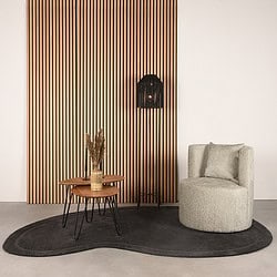 Harig houten wandpaneel met zwart metalen wandlamp, beige fauteuil en salontafel op ronde zwarte vloerkleed in modern interieur.