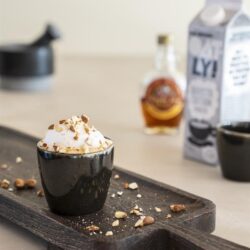 Delicious creamy dessert in black cup topped with nuts on wooden tray with Nutella, milk, and coffee on countertop, cozy kitchen setting, Mokana Meubelen in the background.