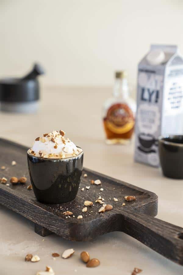 Delicious creamy dessert in black cup topped with nuts on wooden tray with Nutella, milk, and coffee on countertop, cozy kitchen setting, Mokana Meubelen in the background.
