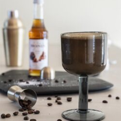 Rijke, romige koffie in een elegant glas met koffiebestanddelen op een witte tafel. Perfect voor koffieliefhebbers die genieten van stijlvolle koffie-ervaringen.