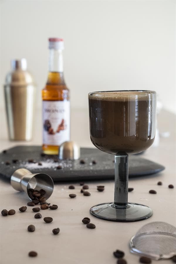 Rijke, romige koffie in een elegant glas met koffiebestanddelen op een witte tafel. Perfect voor koffieliefhebbers die genieten van stijlvolle koffie-ervaringen.