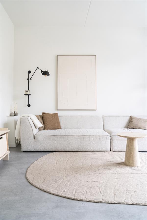 Minimalistisch wit bankstel met zachte beige kussens, moderne zwarte muur lamp en ronde beige tapijt in een strakke, lichte woonkamer van Mokana Meubelen.