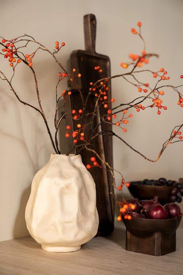 Rustieke witte keramieken vaas met decoratieve takken met oranjerode bessen, vintage houten snijplank en houten kom met uien en rozijnen, stijlvolle interieurdecoratie.