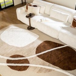 Moderne witte bank met sierkussens en bijzettafel in woonkamer, naaldvilt tapijt met abstracte design, houten vloer en muurdecoratie, stijlvolle woonkamerinrichting.