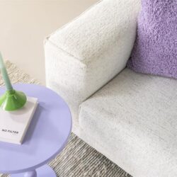 Kleine, elegante woonkamer setup met comfortabel witte bank, paarse sierkussen en een uniek pastelblauw bijzettafeltje met groene kandelaardrab. Perfect voor moderne, gezellige interieurs.