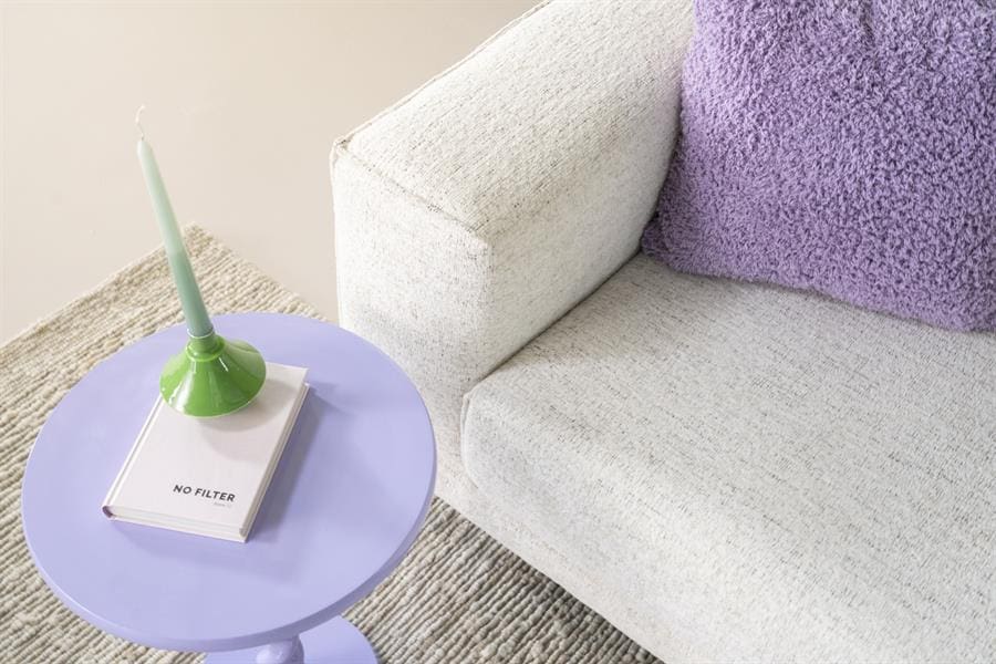 Kleine, elegante woonkamer setup met comfortabel witte bank, paarse sierkussen en een uniek pastelblauw bijzettafeltje met groene kandelaardrab. Perfect voor moderne, gezellige interieurs.