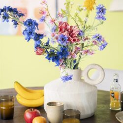 Kunstbloemen in een moderne witte vaas op een eettafel met fruit en ontbijt, stijlvolle interieur decoratie, sfeer van gezelligheid en design, perfect voor een geslaagde woningpresentatie.