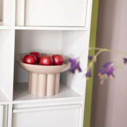 Appel centraal op witte boekenkast met keramische fruitschaal en groene muur voor een modern interieur.