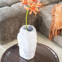 Een witte keramische vaas met een ruwe textuur, met daarin een heldere bloem met oranje tinten, geplaatst op een ronde tafel in een modern woonkamer met een bank en een gebreide deken.