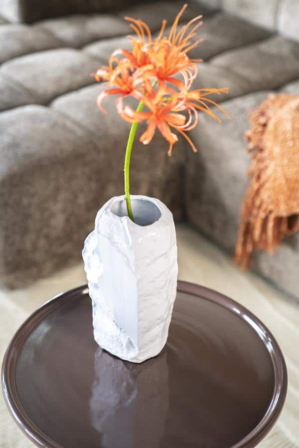 Een witte keramische vaas met een ruwe textuur, met daarin een heldere bloem met oranje tinten, geplaatst op een ronde tafel in een modern woonkamer met een bank en een gebreide deken.