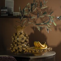 Hoogwaardige keramische vaas met olijftakken en bananen op een houten tafel in een sfeervolle interieur setting.