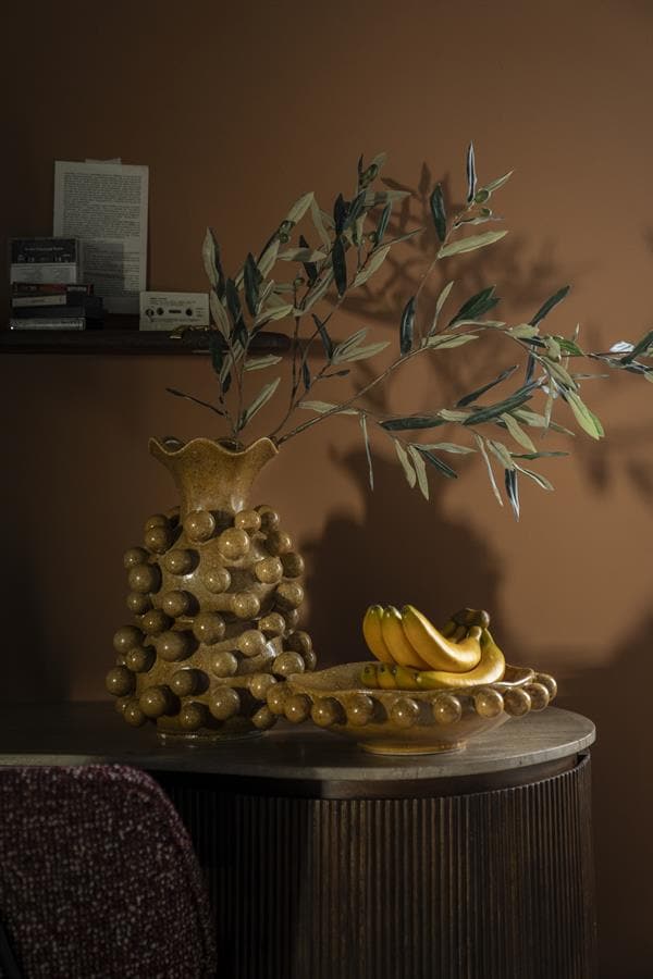 Hoogwaardige keramische vaas met olijftakken en bananen op een houten tafel in een sfeervolle interieur setting.