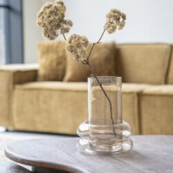 Minimalistische glazen vaas met gedroogde bloemen op een lichte houten tafel in een modern wooninterieur.