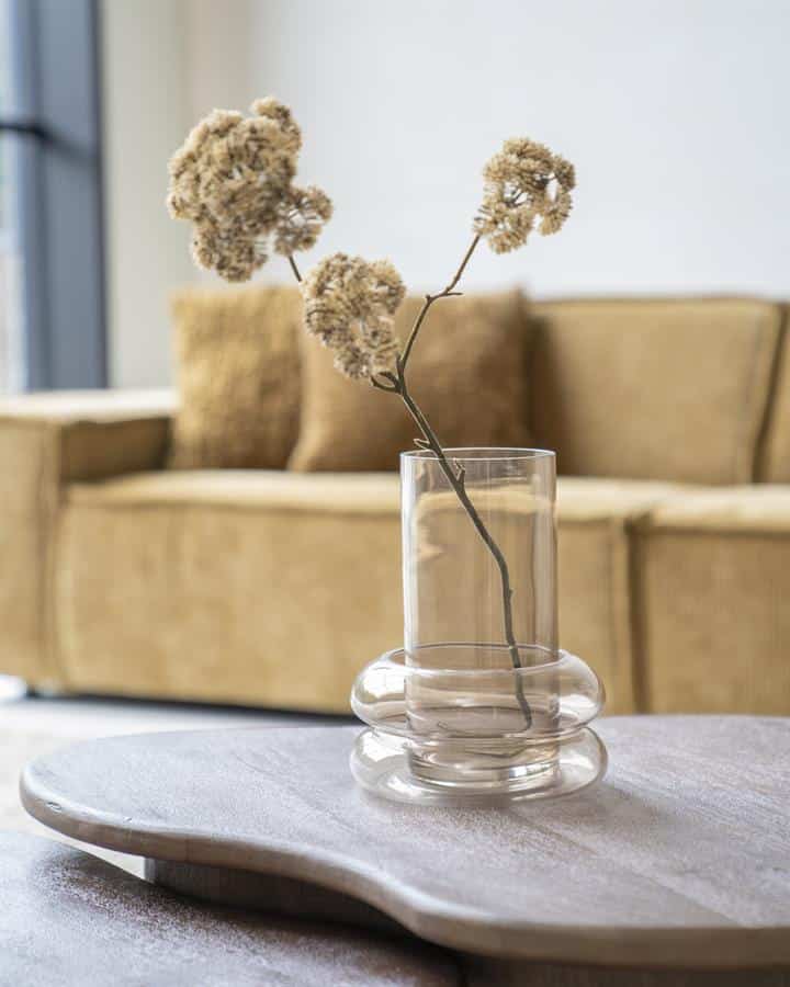 Minimalistische glazen vaas met gedroogde bloemen op een lichte houten tafel in een modern wooninterieur.