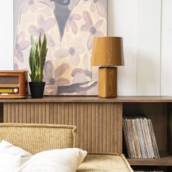 Zandkleurige bank met witte kussens, muurdecoratie met prachtige kunstprint, houten dressoir met vintage radio, groene plant, moderne houten tafellamp, vinyl platen en boeken op plank, stijlvolle woonkamer inrichting, Mokana Meubelen, Scandinavisch interieur.