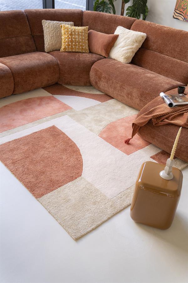 Rustkleurige stoffen hoekbank met sierkussens in neutrale tinten, gestyled in een moderne woonkamer met een abstract geometrisch tapijt, perfect voor gezellige en stijlvolle zithoeken.