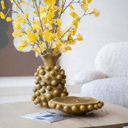 Goudkleurige sierpot met takken met gele bladeren en een bijpassende schaal op een licht houten salontafel in een modern woonkamer.
