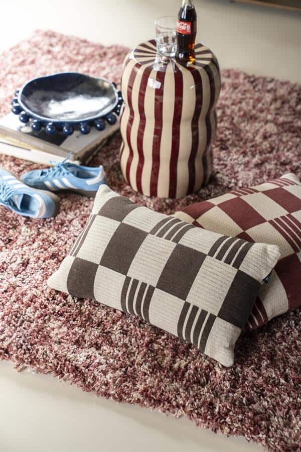 Lange beschrijving: Een gezellige woonstijl met een gestreepte en geblokte sierkussen op een zachte roze/rood vloerkleed met diverse woonaccessoires zoals een glazen fles, een blauwe schoenen, een vaas en een dienblad met een kopje en een fles cola.