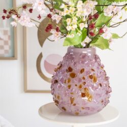 Kunststof vaas met roze en goudkleurige details gevuld met een bos van roze bloemen op een witte bijzettafel.