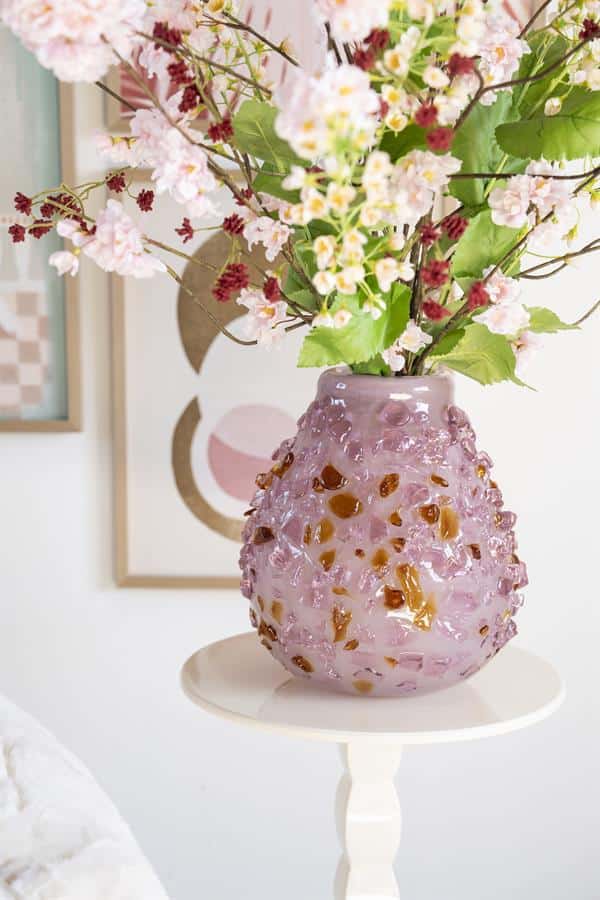 Kunststof vaas met roze en goudkleurige details gevuld met een bos van roze bloemen op een witte bijzettafel.