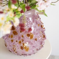 Brocante vaas met roze glas en amber accenten, gevuld met decoratieve bloemen, op een wit bijzettafeltje.