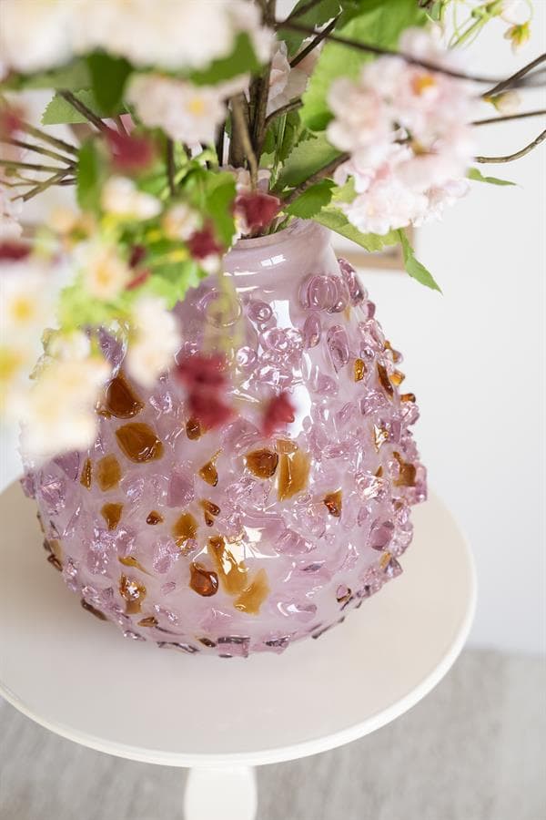 Brocante vaas met roze glas en amber accenten, gevuld met decoratieve bloemen, op een wit bijzettafeltje.