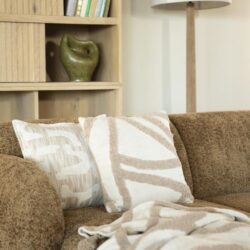 Donkerbruin stoffen bank met beige en witte gestreepte kussens en plaid, houten boekenkast met groene vaas en boeken, moderne woonkamer inrichting, Mokana Meubelen, interieur design.