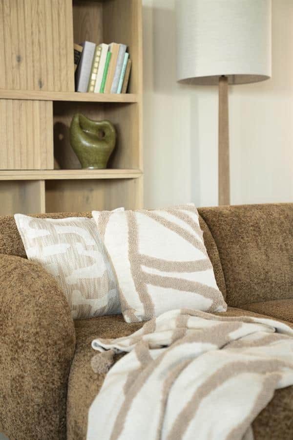 Donkerbruin stoffen bank met beige en witte gestreepte kussens en plaid, houten boekenkast met groene vaas en boeken, moderne woonkamer inrichting, Mokana Meubelen, interieur design.