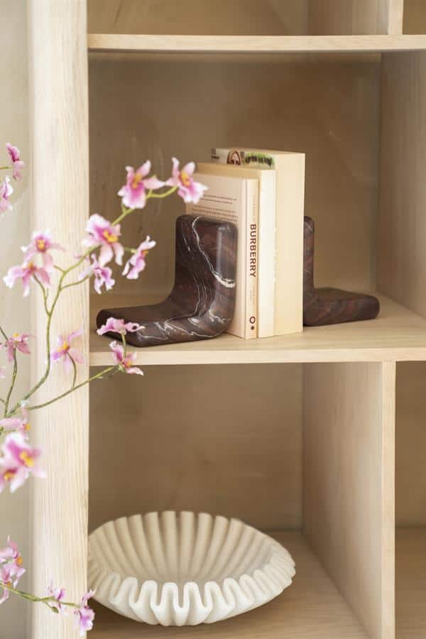 Marmeren schoenenbeeldjes op houten boekenkast gecombineerd met boeken en witte vaas, elegante decoratie voor interieur, stijlvolle meubelaccessoires.