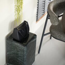 Strakke zwarte leren tas op een groene marmeren bijzettafel naast een modern fauteuil, minimalistisch interieur met groen planten en houten jaloezieën.
