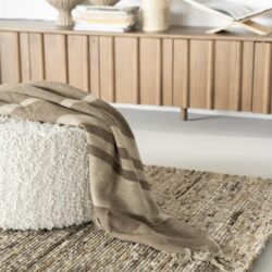Rustieke wollen plaid op een beige stoffen poef met wollen bekleding, gelegen op een jaren 70 stijl laagpolig tapijt, in een modern interieur met een houten tv-meubel en decoratieve vazen op de achtergrond.