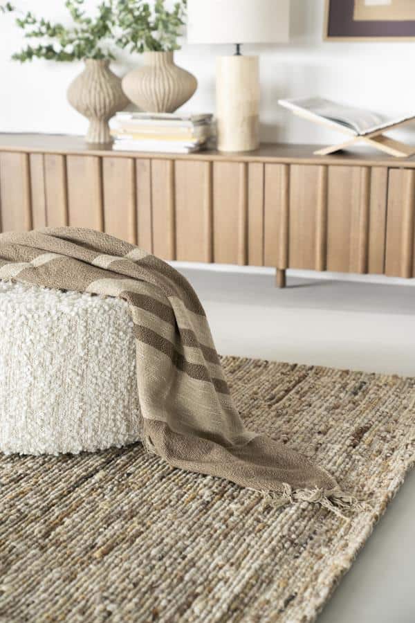Rustieke wollen plaid op een beige stoffen poef met wollen bekleding, gelegen op een jaren 70 stijl laagpolig tapijt, in een modern interieur met een houten tv-meubel en decoratieve vazen op de achtergrond.