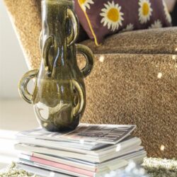 Unieke aardewerken vaas met decoratief ontwerp, geplaatst op een stapel tijdschriften in een gezellige woonkamer met een stoffen fauteuil en bloemenpatroon kussen.