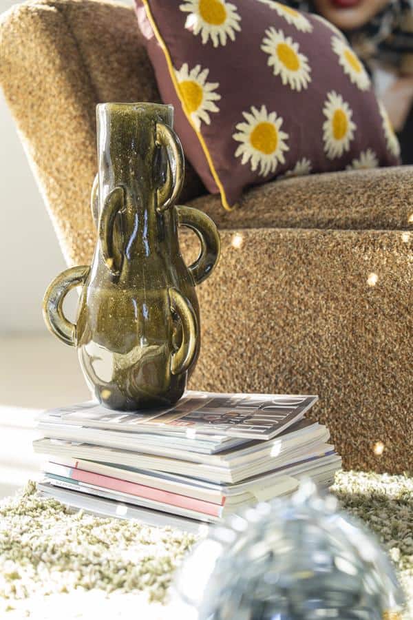 Unieke aardewerken vaas met decoratief ontwerp, geplaatst op een stapel tijdschriften in een gezellige woonkamer met een stoffen fauteuil en bloemenpatroon kussen.