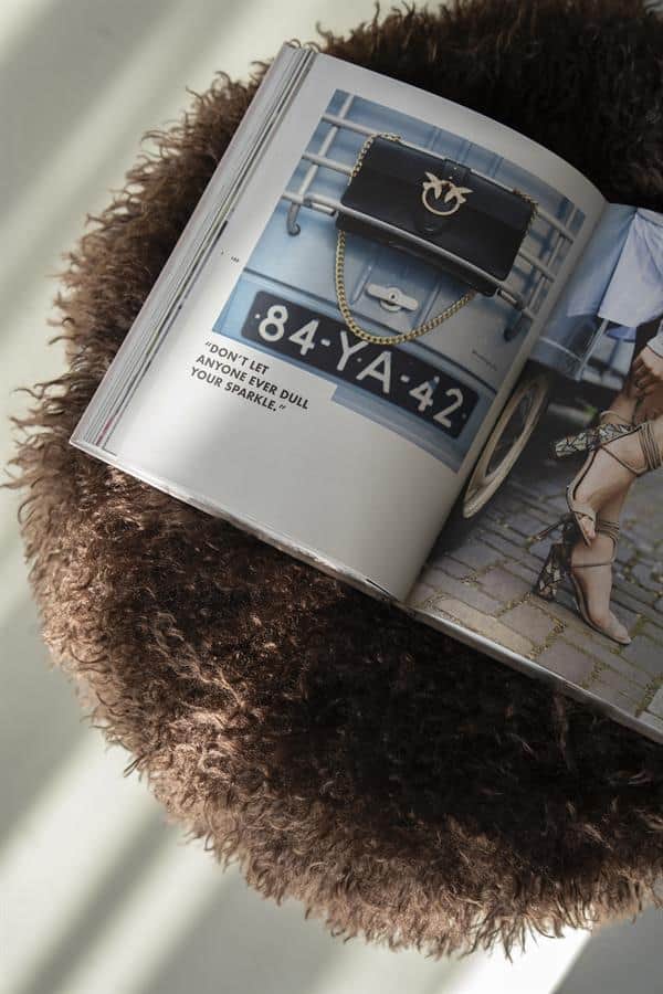 Antiek ogende zwart leren clutch met gouden ketting en logo, op een fotoboek met autoschoolbord numbers en inspirerende quote, gelegen op een schap met een zachte, wollige stoffen schaal, perfect voor stijlvolle interieur en meubeldecoratie.