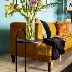 Vaas met frisse bloemen op een zwart bijzettafeltje voor bank in woonkamer met gele bank en kleurrijke kussens.