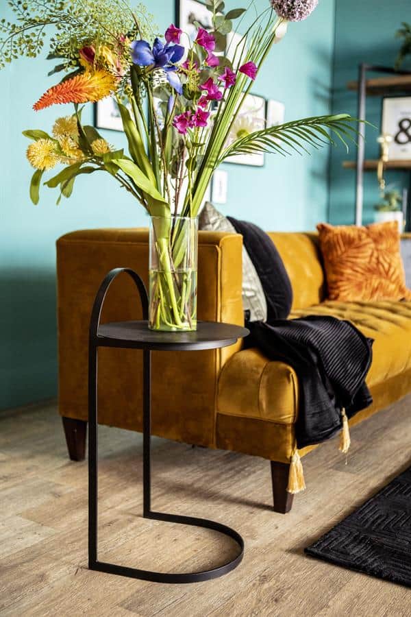 Vaas met frisse bloemen op een zwart bijzettafeltje voor bank in woonkamer met gele bank en kleurrijke kussens.