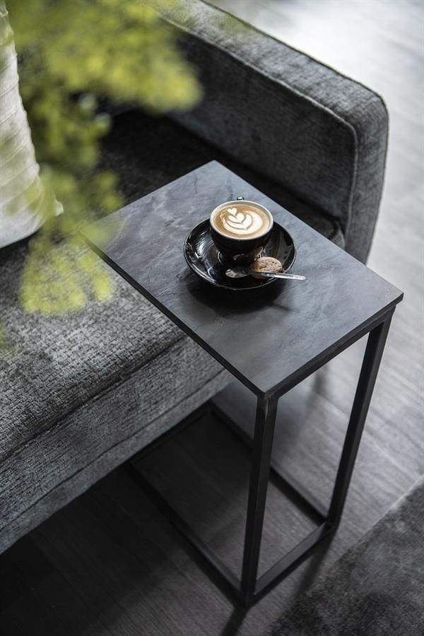 Kleine zwarte bijzettafel naast een comfortabele bank met een kopje koffie en koekje, ideaal voor moderne woonkamers en interieurs.