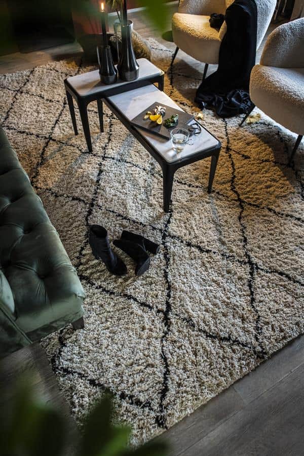 Donkergroene leren bank, zachte beige en grijze fauteuils, glazen salontafel met decoratieve voorwerpen en een modern tapijt met zwart-witte geometrische patronen in een gezellige woonkamer.