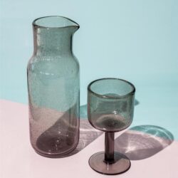 Enkel glasvazen en wijnglas in grijs getint glas, gestyled op een pastelroze en lichtblauwe achtergrond, perfect voor modern interieur en decoratie.